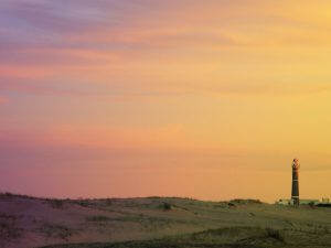 Imagen de un atardecer, en un paisaje de dunas con algo de vegetación; en el cielo aparecen diversos tonos de amarillo, rosados, violetas y naranjas; en el fondo se observa un faro