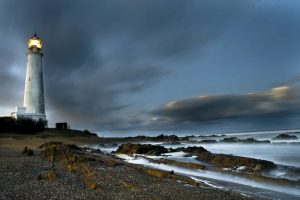Imagen de un faro con su luz encendida en una noche nublada y oscura; a su frente se observan las rocas y parte del Océano Atlántico golpeando contra ellas