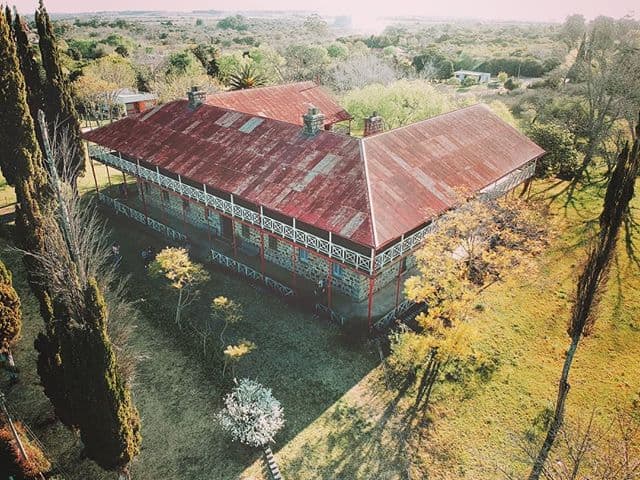 Vista aérea del Hotel Conchillas: un viejo edificio en forma de herradura rectangular, con techos a dos aguas rojo, en medio de un paisaje arbolado