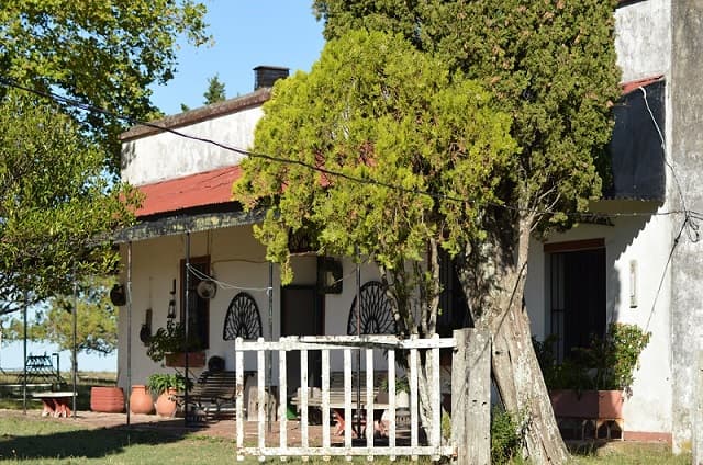 Imagen de un casco de estancia de paredes blancas con un alero de tejas rojo; en la pared hay una vieja rueda de carro y al frente un árbol y un portón de madera blanco