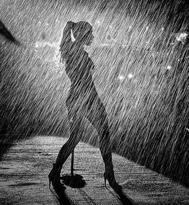 Imagen en blanco y negro desde atrás de una mujer frente a un micrófono de pié, bajo una lluvia intensa
