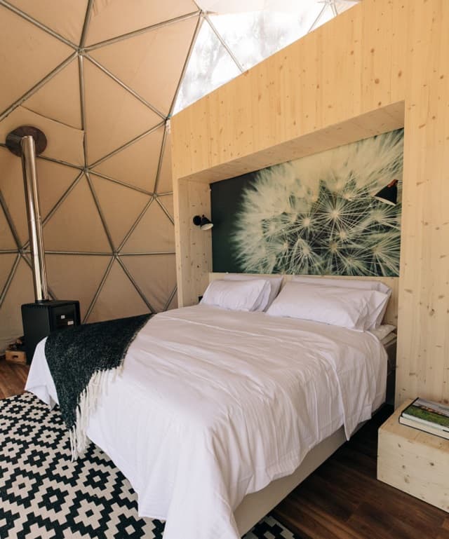 Imagen del interior del domo; se observa un sommier de dos plazas, una pequeña estufa y una alfombra con rombos blancos y negros; arriba, una abertura por donde entra el sol