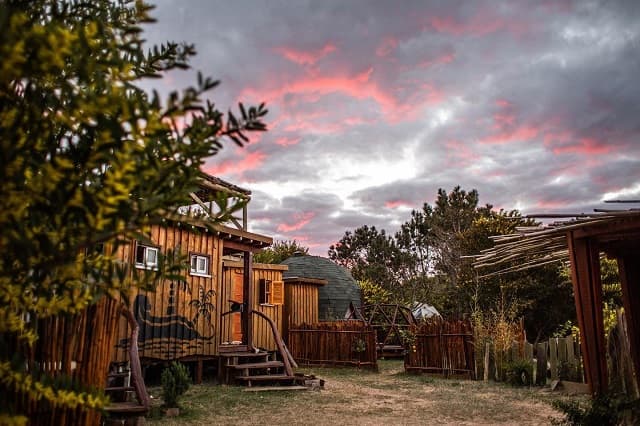 Imagen de varias construcciones de madera, una de ellas en forma de domo,, en medio de la naturaleza. Al fondo se observa el cielo nublado en tonos rosados, naranjas y azules.