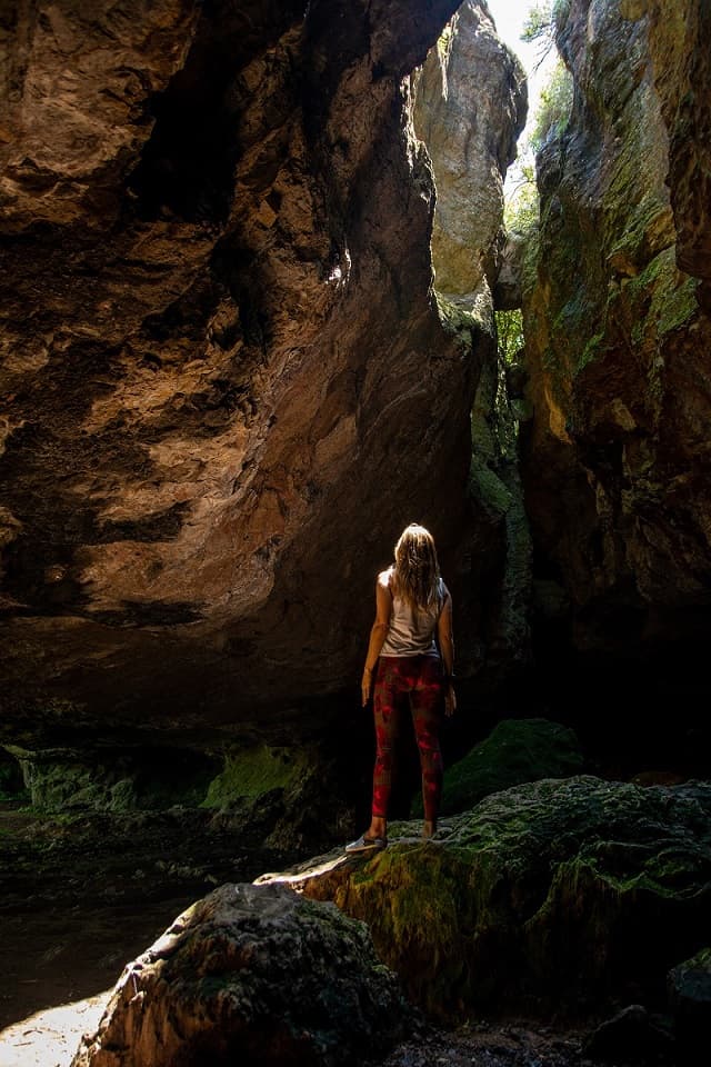 Manuela Da Silveira, parada sobre una gran roca, mirando hacia el espacio abierto por donde entra luz, dentro de una gruta 