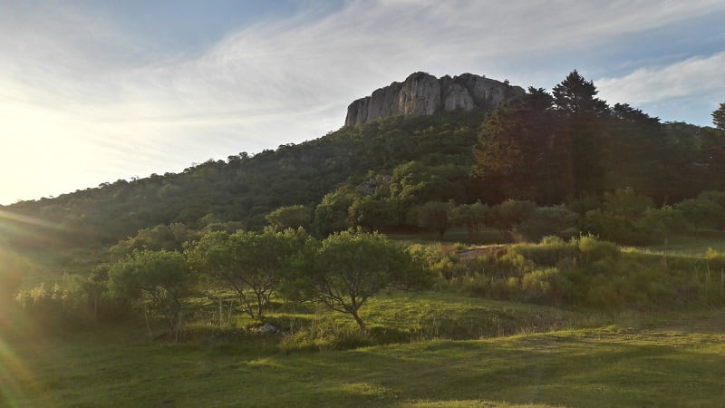 Imagen de un cerro con sus paredes de piedra gris, sobre un paisaje verde, al atardecer