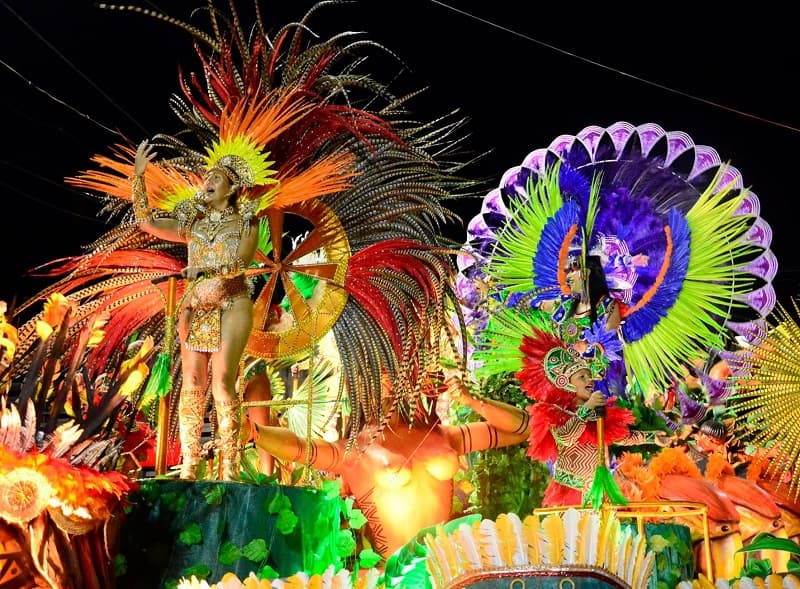 Sobre una corroza llena de colores, varias personas con diferentes disfraces bailan y cantan