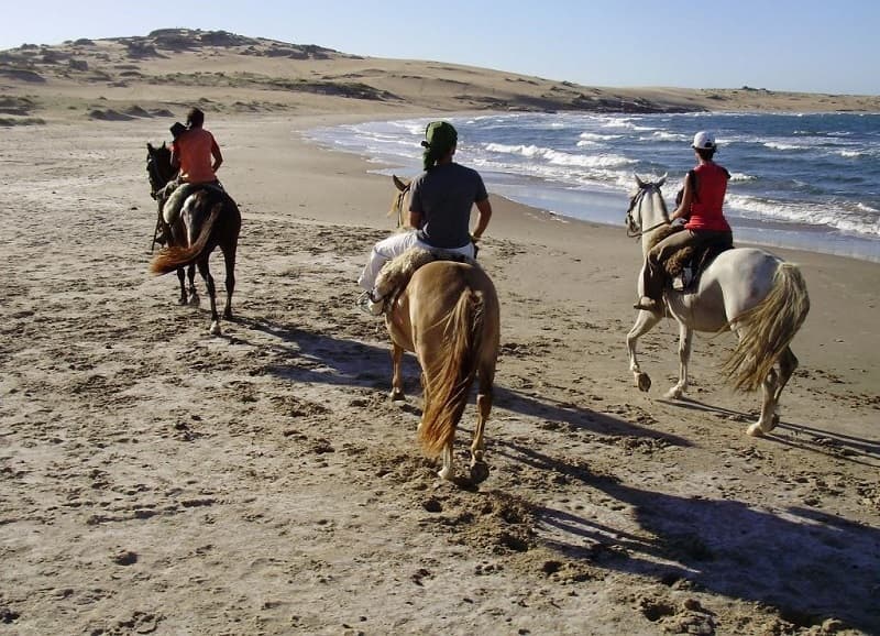 Imagen de tres personas cabalgando sobre la arena, a la orilla del océano; al fondo se observan dunas.