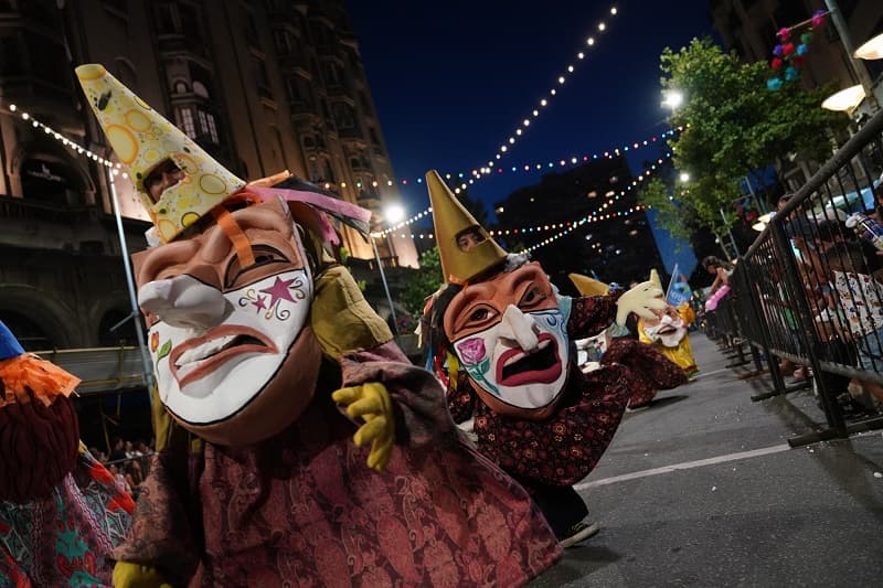 Dos cabezudos con la cara pintada y narices largas desfilan por 18 de julio; sobre la avenida hay guirnaldas de luces de colores.