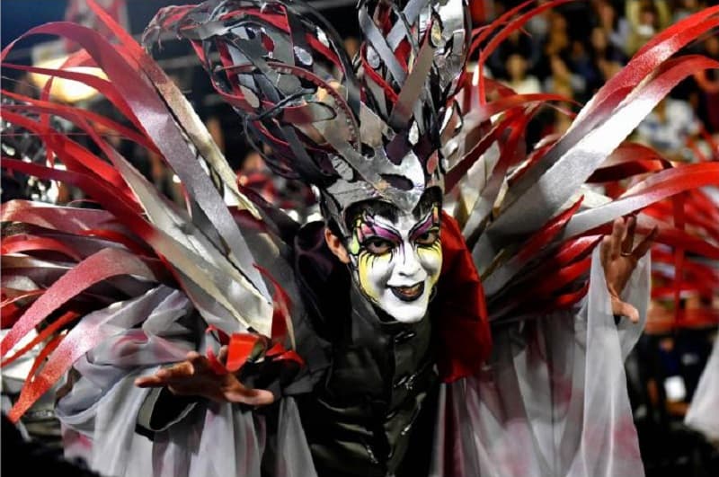 Un hombre con la cara pintada sonríe a la cámara; está vestido con un disfraz y un sonmbrero de formas irregulares de colores rojo, blanco y negro