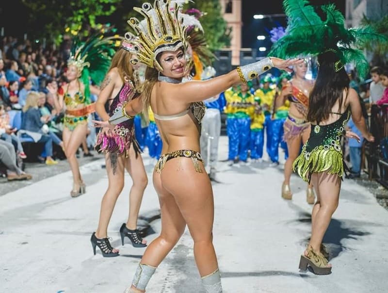 Una mujer con disfraz de carnaval baila y sonríe mientras desfila; detrás se observan más personas desfilando