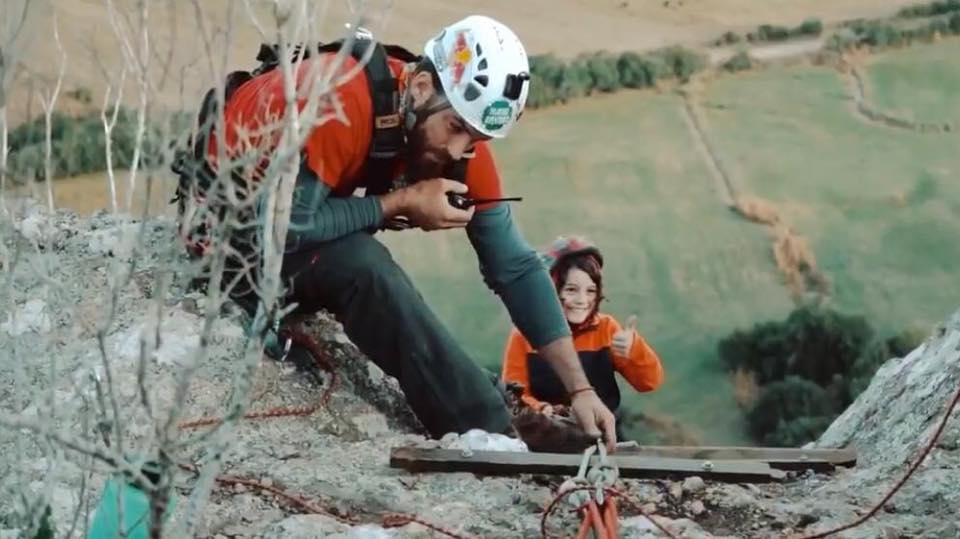 Un niño sonriente comienza el descenso en rapel por una alta pared de piedra, mientras un hombre a pocos metros de él verifica los implementos de seguridad y habla por un equipo de radio