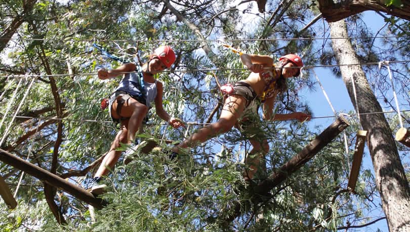 Dos mujeres adolescentes sonrientes caminan sobre cuerdas en entre los árboles