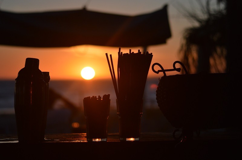 Imagen del sol cayendo sobre el océano; delante hay una coctelera y dos vasos con tragos