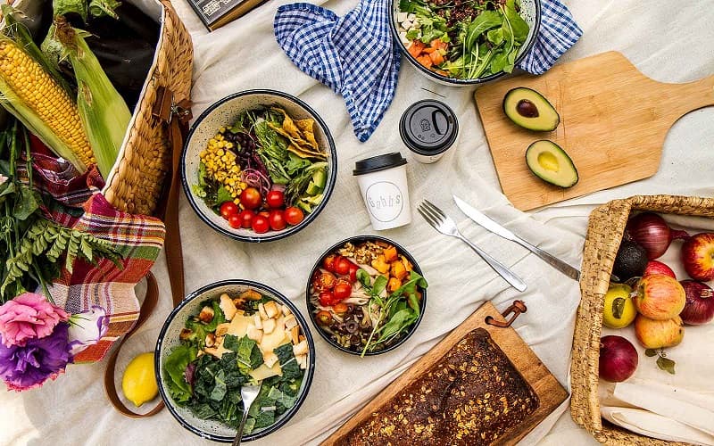 En una mesa, varios bowls con ensaladas distintas, alrededor de las cuales hay tablas, verduras, y panes