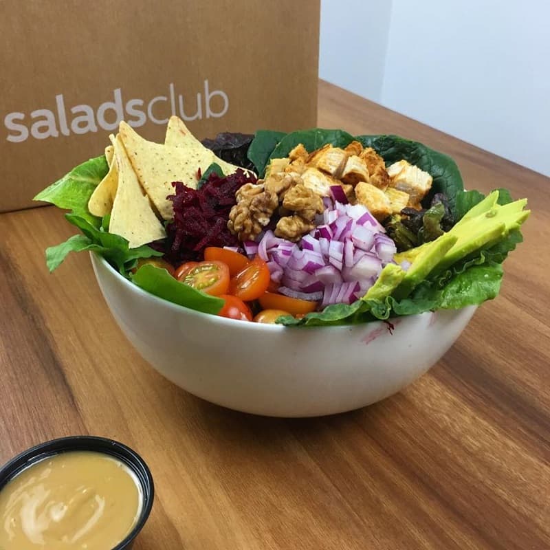 Un bowl blanco con ensalada multicolor y un pequeño pote de salsa; detrás, una bolsa de papel que dice Salads Club