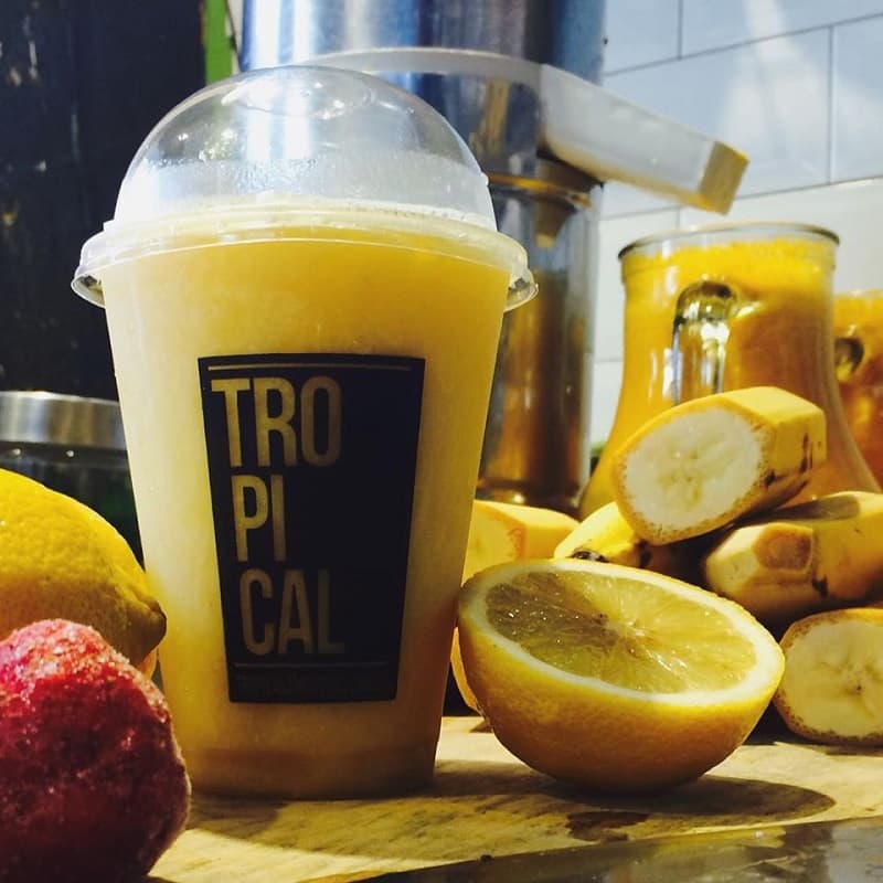 Un vaso plástico con un licuado de frutas amarillas, rodeado de diversas frutas