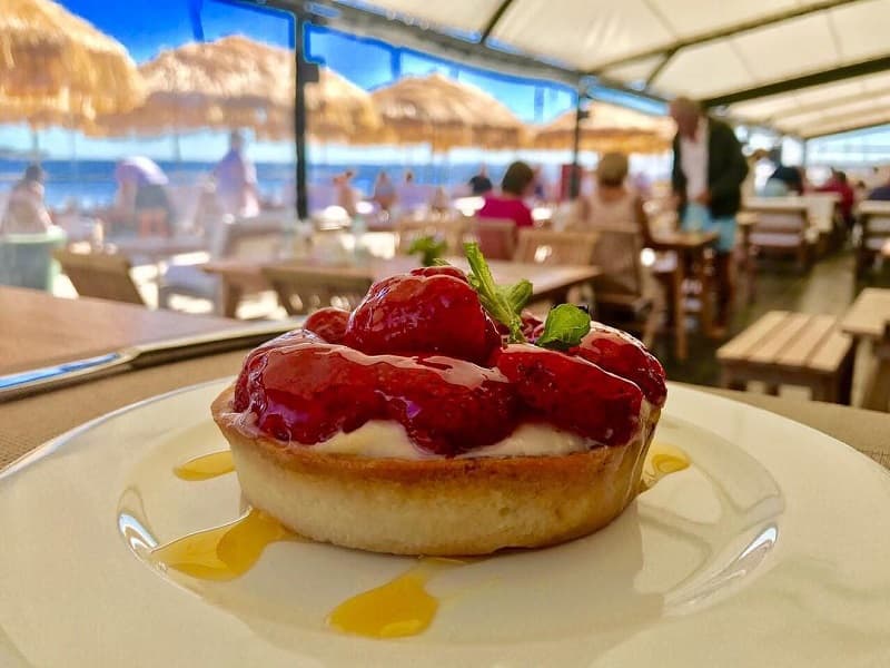 Imagen de una cheesecake de frutilla, sobre un plato blanco; al fondo se observan mesas y personas almorzando en restaurante de Punta del Este donde ofrecen descuentos con tarjetas