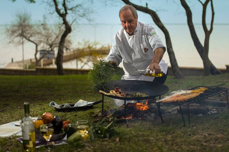 Un cheff prepara un plato sobre una gran sartén en un espacio abierto frente al Río Uruguay
