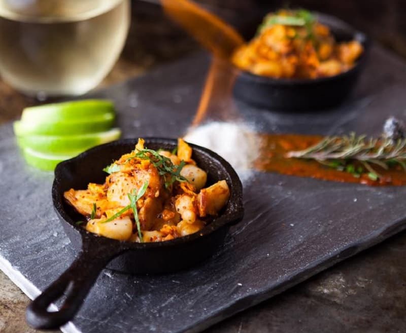 Imagen de una sarten negra con papas rústicas, sobre una tabla gris; a un costado, una copa de vino blanco, en restaurante de Punta del Este donde ofrecen descuentos con tarjetas