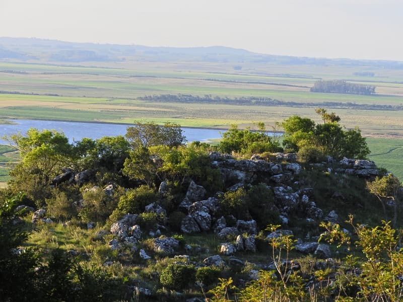 Vista desde la cima de un cerro de un paisaje de sierras verde, con un lago al centro, en Paso Centurión, uno de los parques nacionales de Uruguay