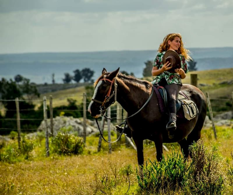 Una mujer cabalgando, sosteniendo un sombrero de ala entre sus manos; al fondo, un paisaje de sierras