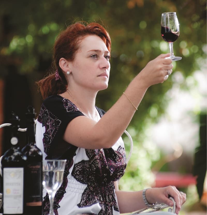 Una mujer levanta una copa con vino tinto; a su costado hay una botella de vino y una copa de agua