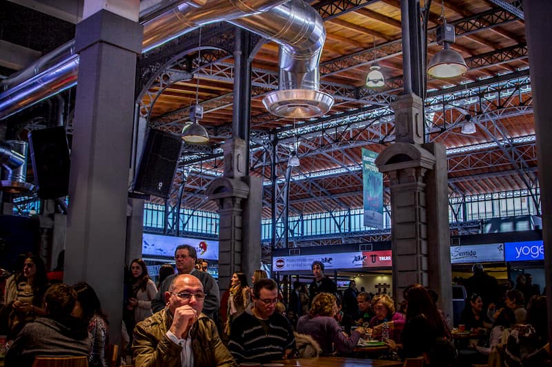 Varias personas cenando en mesas, dentto de un espacio de techos muy altos con ventanales y estructuras de hierro