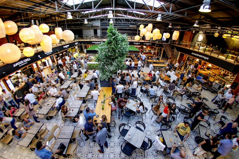 Muchas personas sentadas en un gran espacio cerrado; en el techo hay luces envueltas en globos blancos de papel