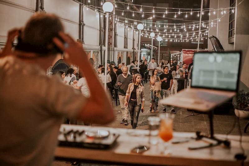 Varias personas caminan y otras están sentadas en mesas, sobre un callejón, frente a un DJ que pone música